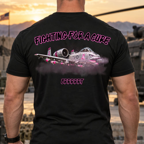 A man wearing a black t-shirt with a back graphic of a pink and gray A10 Warthog fighter jet, the text 'FIGHTING FOR A CURE' and 'BRBRRT', set against a blurred military base background at sunset.