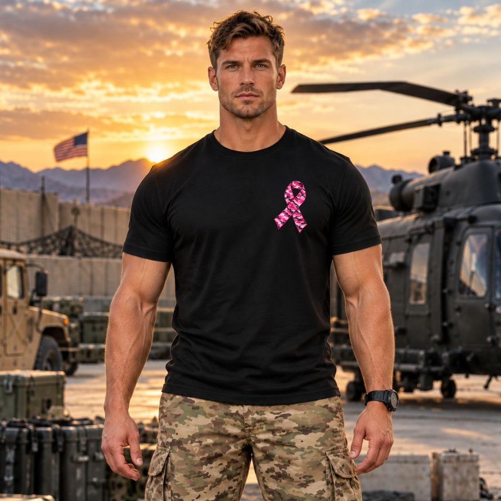 A muscular man wearing a black short-sleeve t-shirt with a pink ribbon graphic on the chest, standing in a military setting with a helicopter and mountains at sunset.