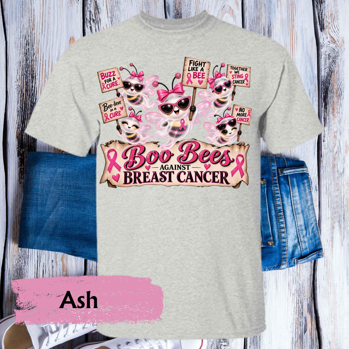 An ash heather gray graphic t-shirt featuring cartoon bees wearing sunglasses and pink bows, holding signs about breast cancer awareness, with pink ribbons and the text 'Boo Bees AGAINST BREAST CANCER'. The shirt is styled flat on a rustic white wood background with blue denim shorts and a pink color label overlay reading 'Ash'.