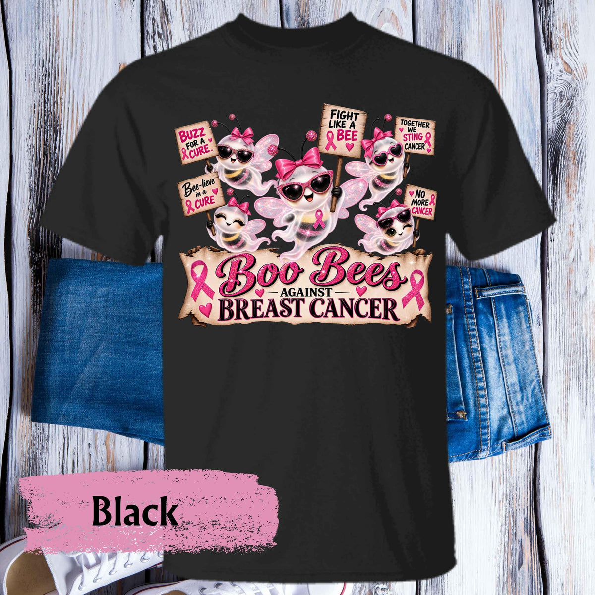 A black graphic t-shirt featuring a 'Boo Bees Against Breast Cancer' design with cartoon bees, pink ribbons, and protest signs, styled on a white wood plank background with blue jeans and a pink 'Black' color label overlay.
