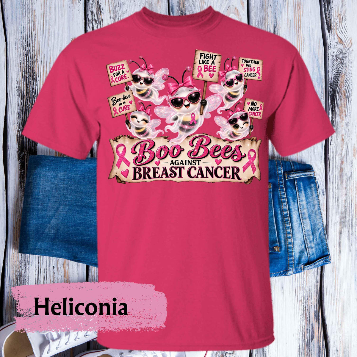 A bright pink graphic t-shirt featuring cartoon bees wearing sunglasses and breast cancer awareness ribbons, with the text 'Boo Bees AGAINST BREAST CANCER'. The shirt is displayed flat on a whitewashed wood background with blue denim shorts behind it.