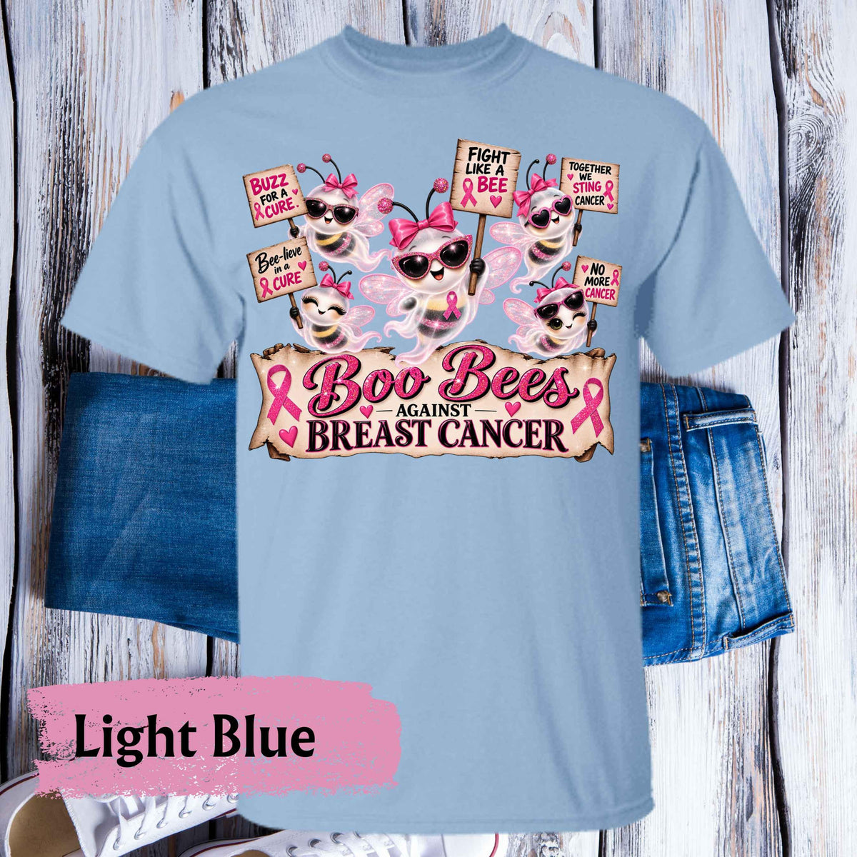 A light blue graphic t-shirt featuring cartoon bees wearing breast cancer awareness sunglasses and pink bows, with signs reading 'Fight Like A Bee' and 'Boo Bees AGAINST BREAST CANCER'. The shirt is displayed flat on a whitewashed wood background with blue jeans as styling props, and a pink label in the corner reads 'Light Blue'.