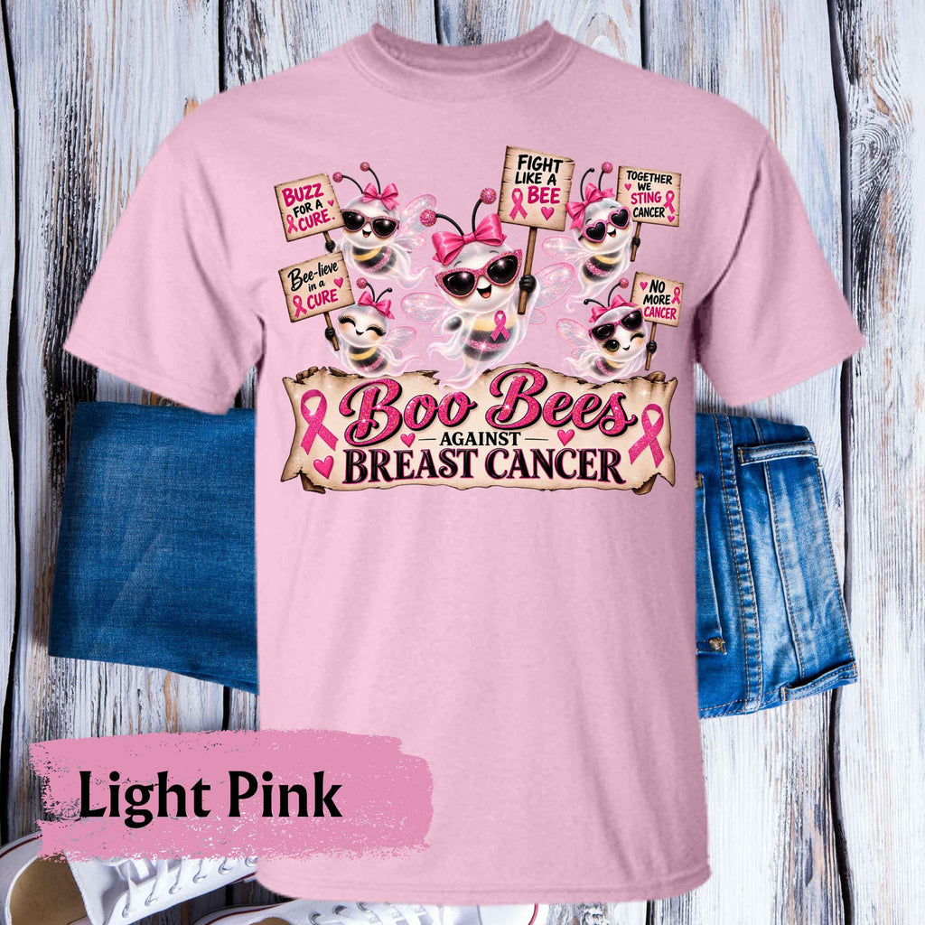 A light pink graphic t-shirt displayed on a rustic white-washed wood background, styled with blue denim shorts. The t-shirt features a 'Boo Bees Against Breast Cancer' design with cartoon bees wearing pink sunglasses and holding breast cancer awareness signs.