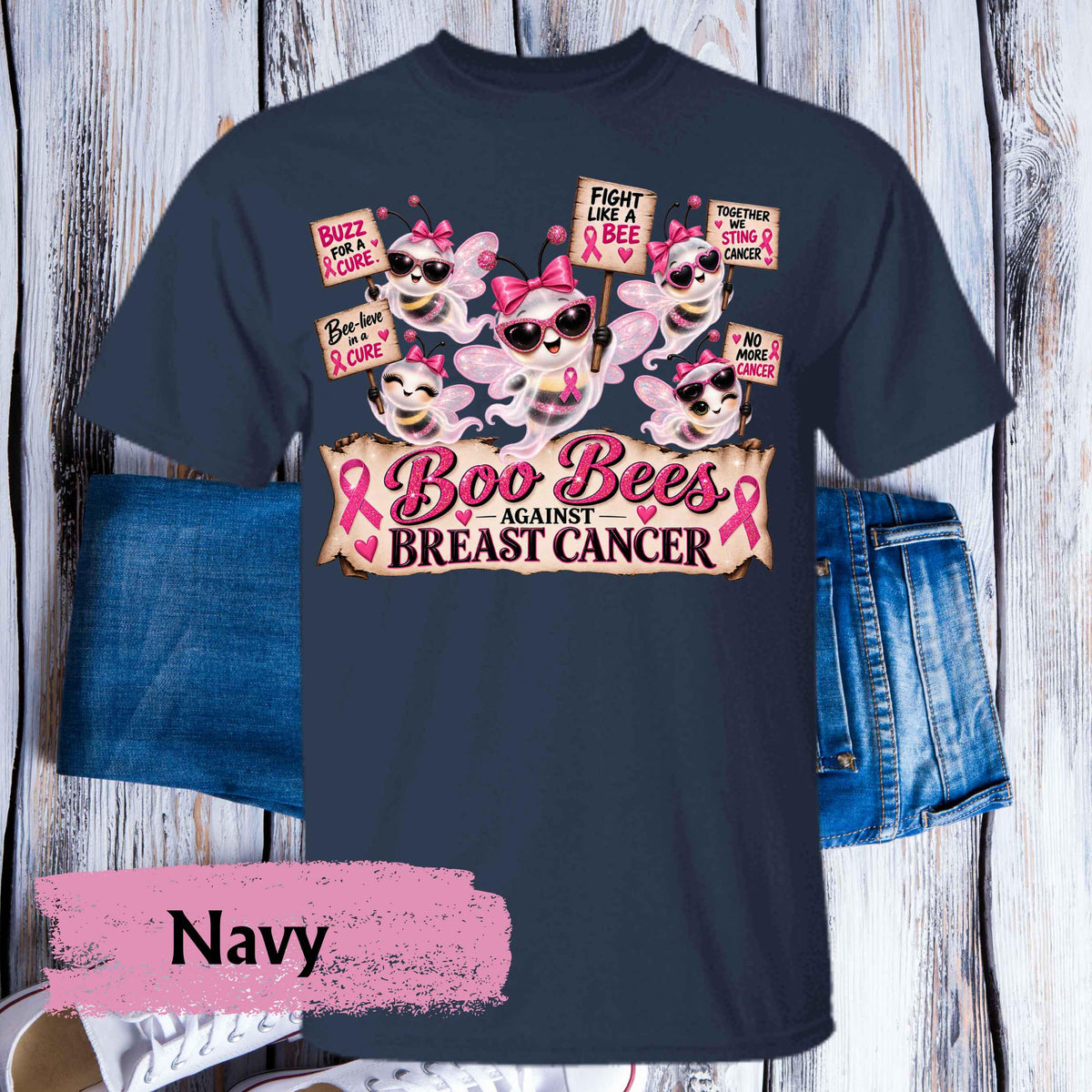 A navy blue graphic t-shirt featuring cartoon bees wearing pink sunglasses and bows, with signs reading 'FIGHT LIKE A BEE' and 'BOO BEES AGAINST BREAST CANCER,' styled with folded denim shorts on a whitewashed wood background. A pink brushstroke label at the bottom reads 'Navy.