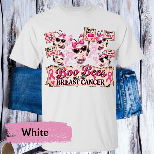 A white graphic T-shirt laid flat on a rustic whitewashed wood background, styled with blue denim shorts behind it. The shirt features a breast cancer awareness design with cartoon bees wearing sunglasses and pink bows, pink awareness ribbons, and the text 'Boo Bees AGAINST BREAST CANCER.