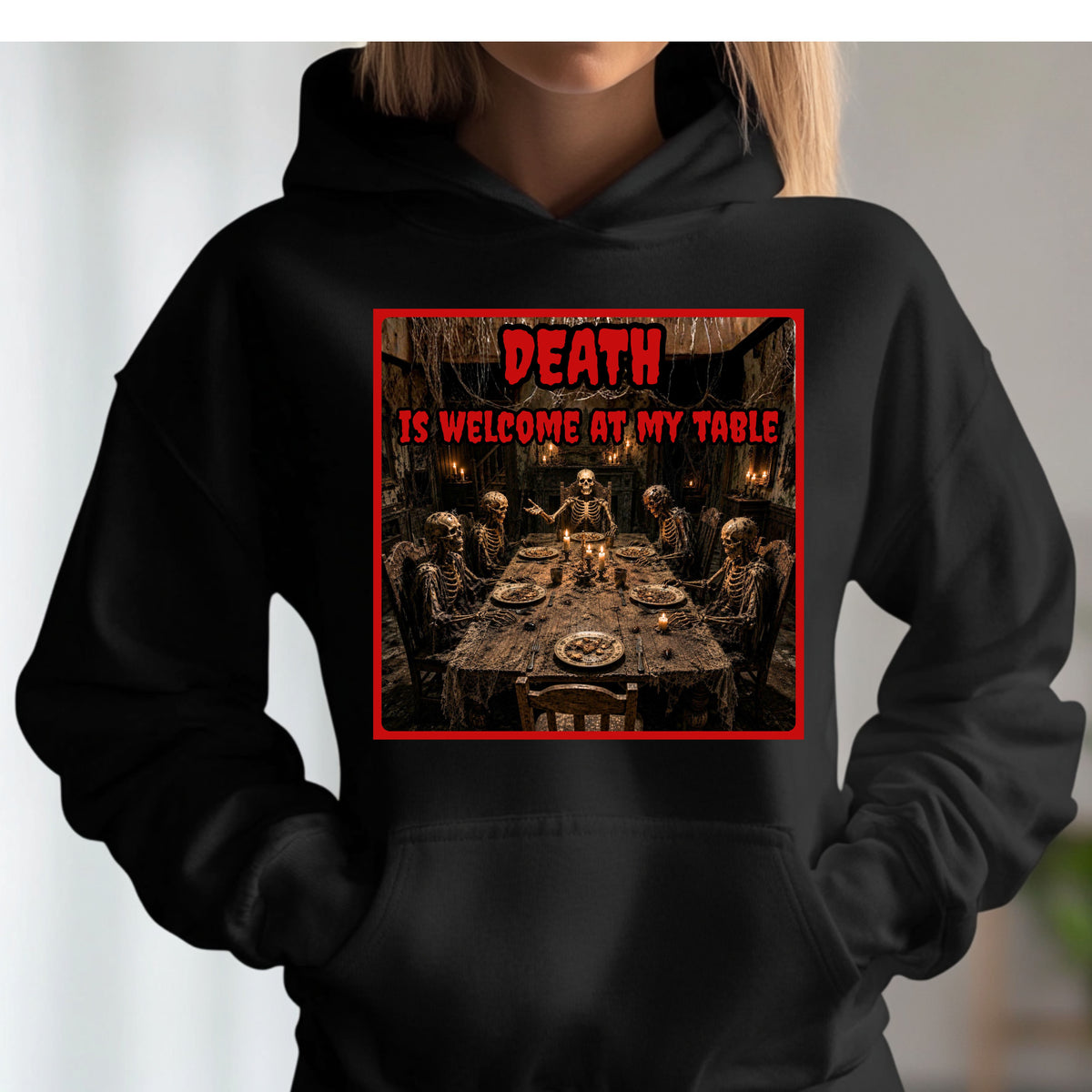 A person wearing a black hoodie with a graphic print on the chest. The print features a dark, gothic scene of skeletons gathered around a table, framed by a red border with the text 'DEATH IS WELCOME AT MY TABLE' in red and black.