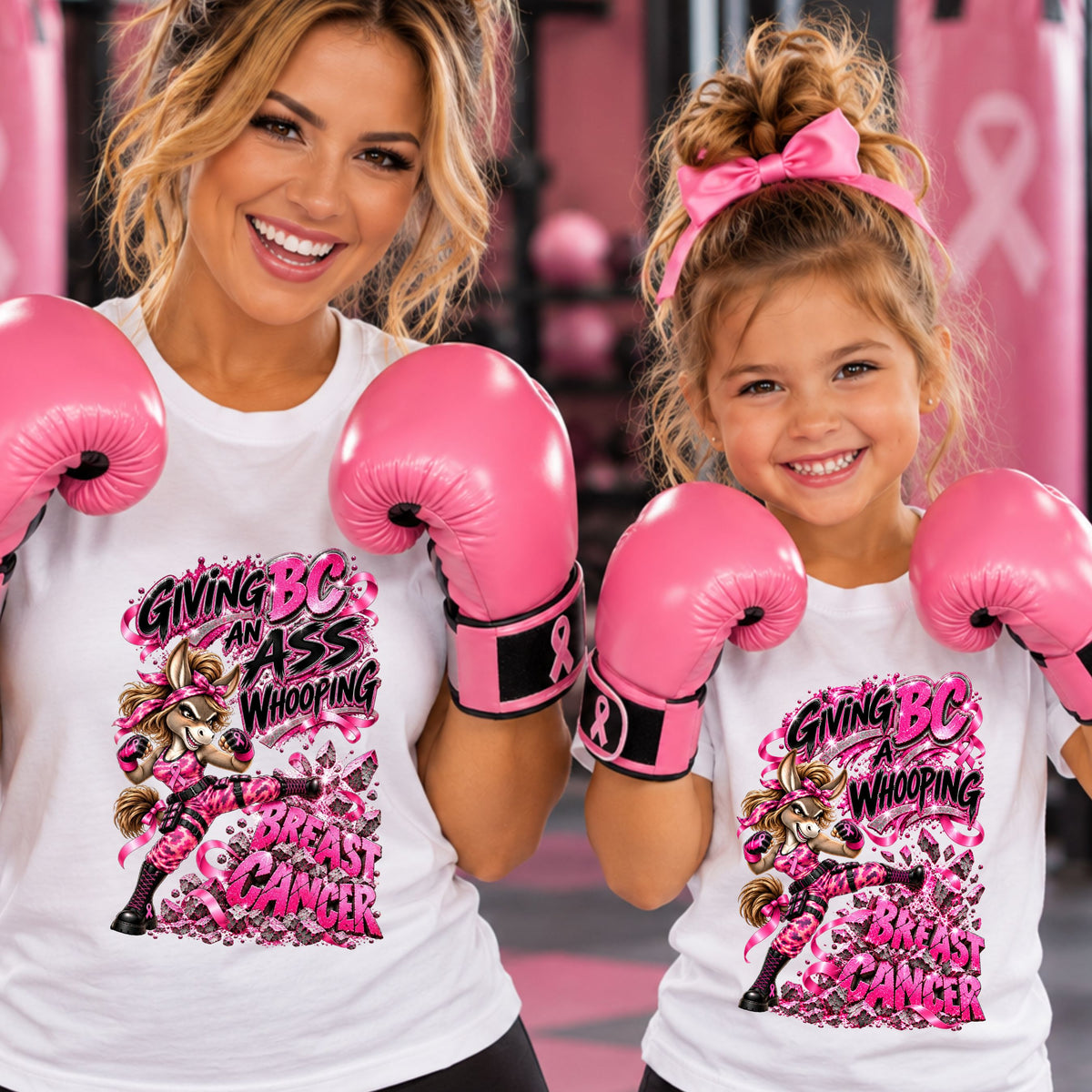A smiling woman and a young girl wearing matching white graphic T-shirts and pink boxing gloves in a pink-themed setting. The shirt features a bold graphic of a fierce donkey kicking the words BREAST CANCER, with the text 'GIVING BS THE ASS WHOOPING" for the woman's shirt, and "GIVING BS A WHOPPING" for the young girl's shirt.