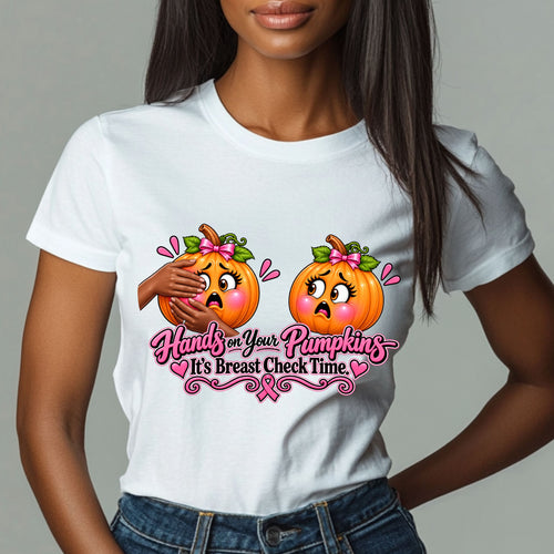 A woman wearing a white short-sleeve graphic T-shirt with a playful Breast Cancer Awareness Halloween design featuring two adorably horrified pumpkins, pink bows, pink accents, a breast cancer awareness ribbon and the cheeky reminder: “Hands on Your Pumpkins — It's Breast Check Time!”