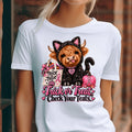 A woman wearing a white short-sleeve graphic T-shirt. The shirt features a cartoon Highland cow in a black cat costume holding a pink 'Trick or Treat' bag next to a pink pumpkin, with the text 'Trick or Treat Check Your Teats.