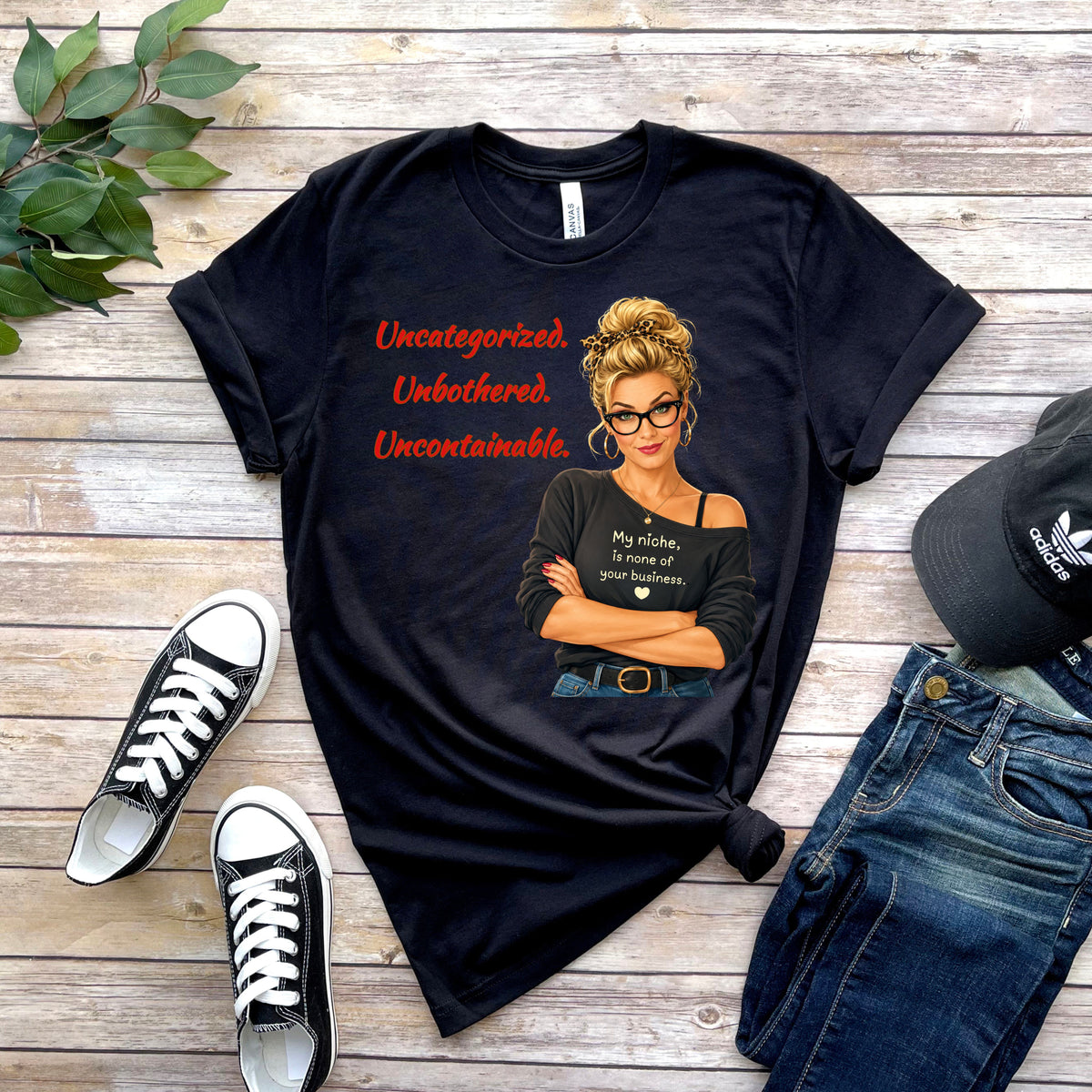 A black graphic t-shirt laid flat on a rustic wood background, styled with black sneakers, blue jeans, and a black Adidas cap. The t-shirt features a graphic of a woman with glasses and text that reads “Uncategorized. Unbothered. Uncontainable.” with the perfectly cheeky declaration:

“My niche is none of your business.”
