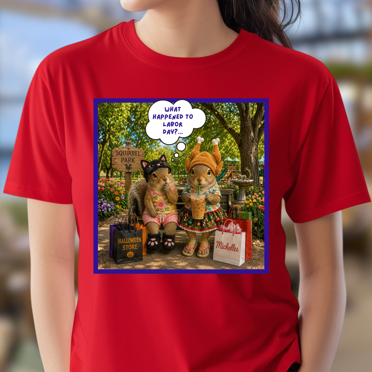 A person wearing a bright red short-sleeve T-shirt with a large square graphic print on the chest. The print shows two fashionable squirrel besties are taking a well-deserved shopping break at Squirrel Park. One is clearly ready for spooky season in her cat costume, while the other has apparently skipped straight past fall and landed somewhere between Thanksgiving and Christmas—iced coffee in hand, naturally. Surrounded by shopping bags and seasonal chaos, one squirrel wonders "What Happened to Labor Day?"