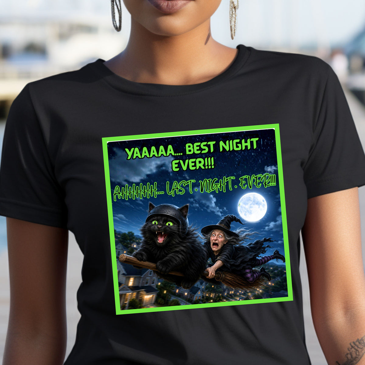 A woman wearing a black short-sleeve t-shirt with a colorful graphic print on the front. This Halloween design features a wildly enthusiastic fluffy black cat—helmet on, green eyes glowing, and having the absolute time of his nine lives—as he rockets through the night sky on a witch’s broom. Hanging on behind him is one completely terrified witch who looks like she’d like to get off this ride immediately. The bold neon-green lettering has the cat screaming: “YAAAAA… BEST NIGHT EVER!!!”.