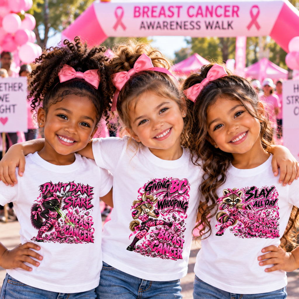 Three young girls wearing matching white graphic T-shirts with pink and black designs, standing together at a Breast Cancer Awareness Walk under a pink ribbon arch. The girl on the left is wearing a shirt with a black cat staring down breast cancer, the middle girl is wearing a white shirt which has a donkey kicking breast cancer, and the girl on the right has a white shirt with a raccoon slaying breast cancer.